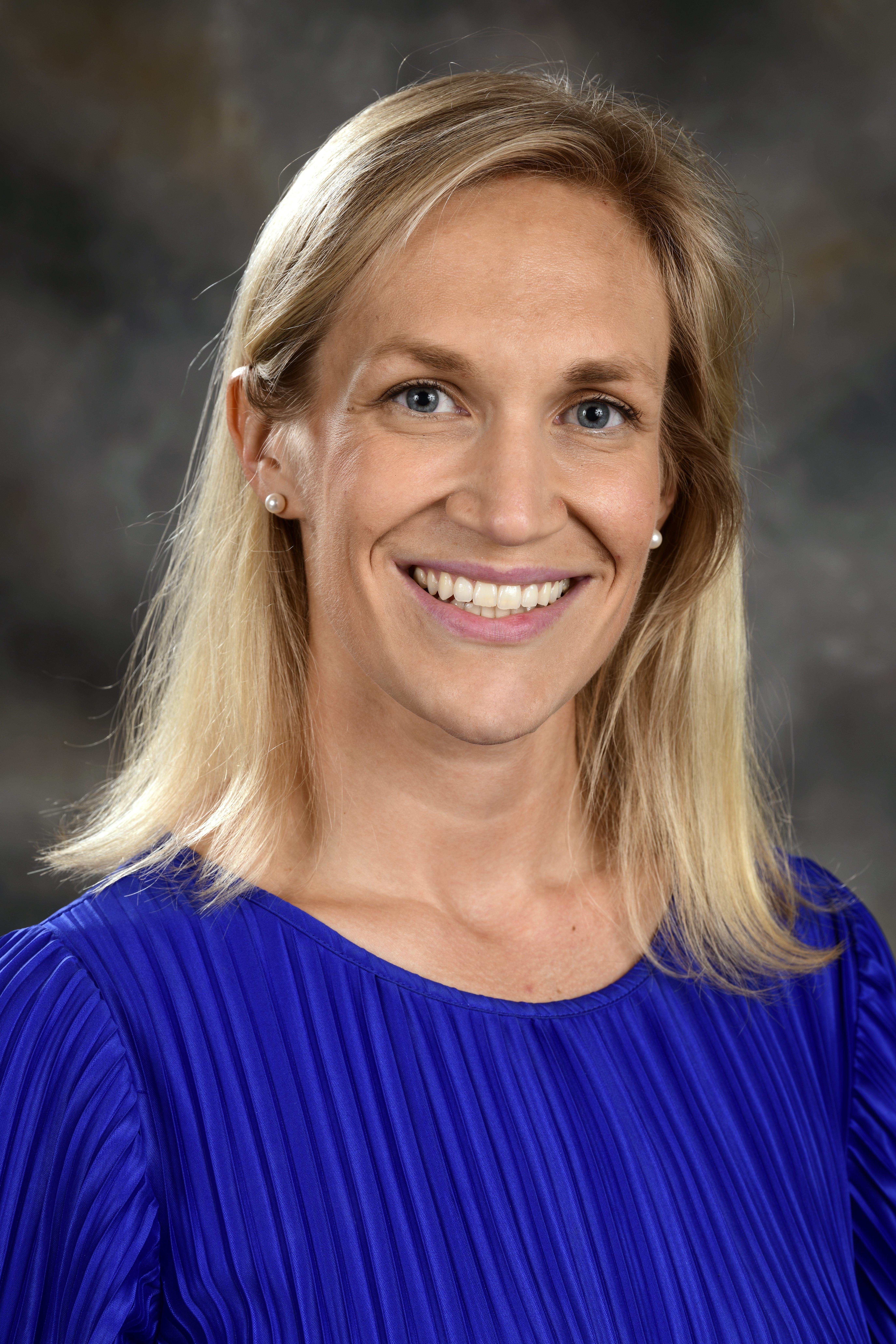 Headshot of a white woman with shoulder-length blonde hair, blue eyes, wearing a royal blue dress against a gray background